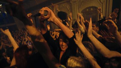 A crowd of people dancing together in a dimly lit ornate room with arms in the air