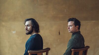 Two men sat in wooden chairs next to each other, looking over their left shoulders
