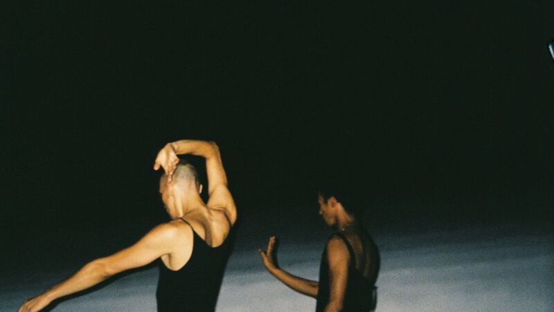 Two people stood toward a dark space and their back to the camera wearing black tops. Their arms are lifted in different positions