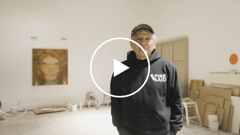 Artist Yoshitomo Nara – an older Japanese man with grey hair over which he wears a black baseball cap, and wearing a black hooded top, stands in his whitewalled artists studio which has paintings on the wall and various materials propped against the walls and at the room's edges.