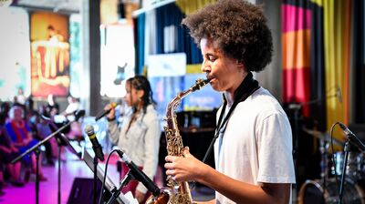 A young person playing the saxophone with someone singing into a mic behind them