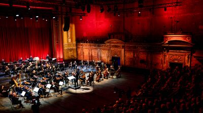Concert Orchestra on stage under stage lighting in a large auditorium with wooden floors