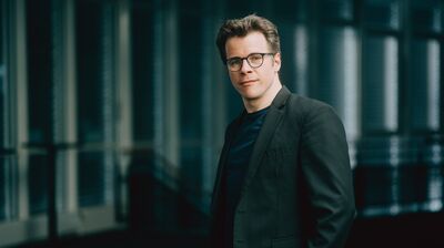 A man with brown hair and glasses in a dark suit looking into the camera with large glass windows behind