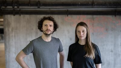 A man and woman in neutral t shirts, stood in front of a concrete wall