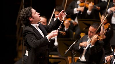 Conductor wearing a black suit white shirt and pink bow tie, with violinists playing around them