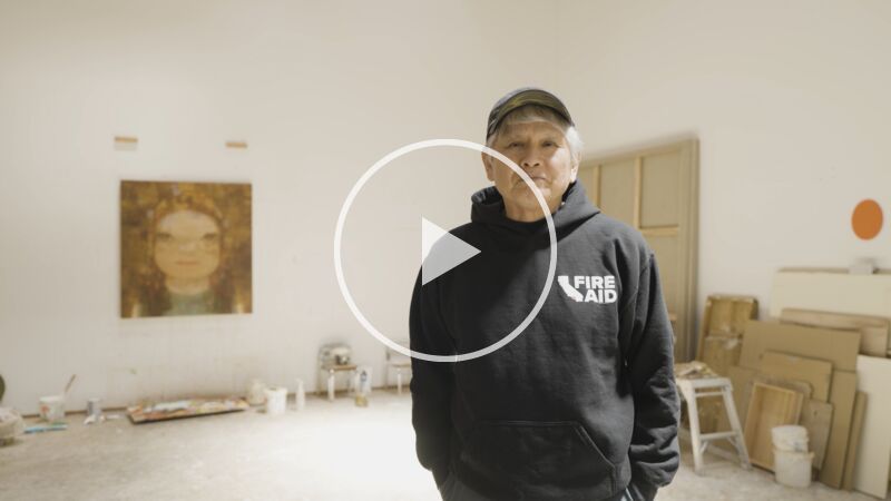 Artist Yoshitomo Nara – an older Japanese man with grey hair over which he wears a black baseball cap, and wearing a black hooded top, stands in his whitewalled artists studio which has paintings on the wall and various materials propped against the walls and at the room's edges.