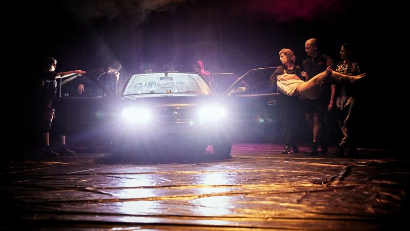 A car with its headlights on and doors open, three people are carrying someone wearing white