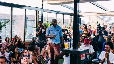 A person in a blue shirt moving through a seated crowd who are clapping. They are being filmed and in a large open air space.