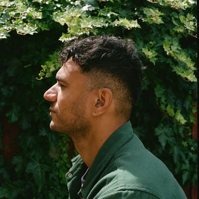 Raymond Antrobus sitting sideways on wearing a green jacket against a leafy background.