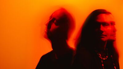 Two faded and blurry figures wearing black against an orange background
