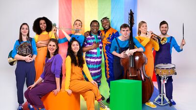 A group of musicians pose together in front of a vibrant rainbow-colored backdrop. They are dressed in bright colours, and are holding various instruments