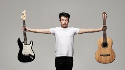 Person wearing a white t-shirt holding an electric guitar in his left hand and an acoustic guitar in his right at arms length