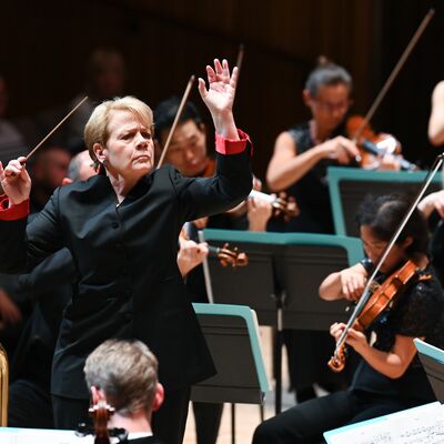 A conductor holding a baton in one hand raising both arms signalling to a group of seated violinists