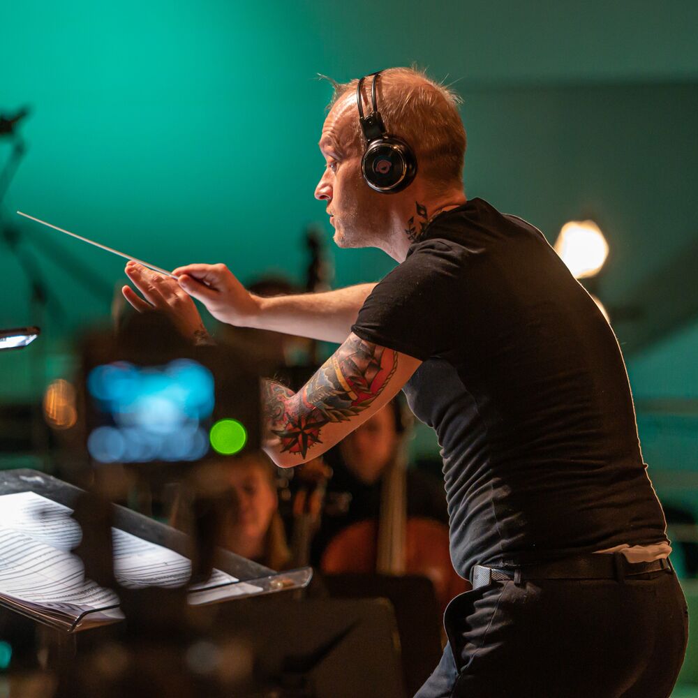 Man with tattoos conducting a classical ensemble in a green room, wearing headphones, black pants and a black t-shirt
