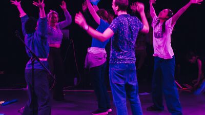 Group of people in a room dancing with their arms in the air lit in purple lighting