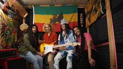 A group of four musicians smile at the camera holding an electric guitar with a Rastafarian flag behind them
