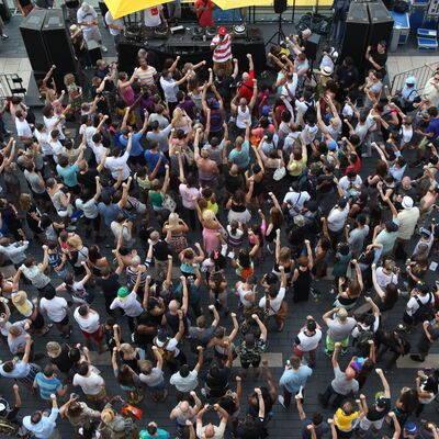 Ariel view of a crowd surrounding dj decks and speakers with one MC in a striped top, people are lifting one arm up