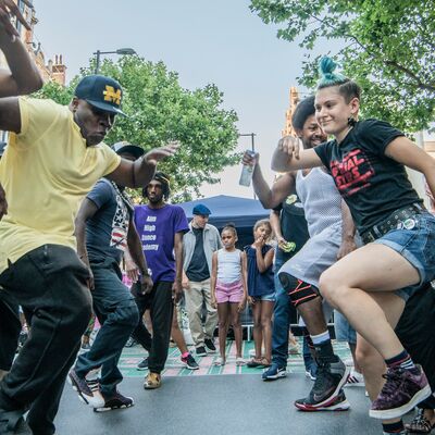 A crowd dancers together on a street with buildings and trees in the background