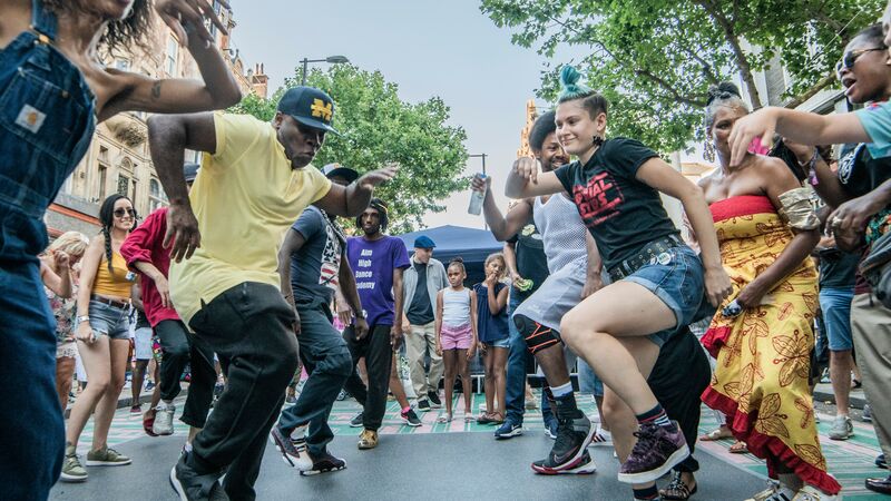 A crowd dancers together on a street with buildings and trees in the background