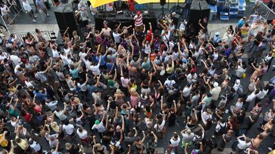 Ariel view of a crowd surrounding dj decks and speakers with one MC in a striped top, people are lifting one arm up