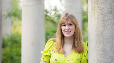 Elodie Harper wearing a yellow dress standing against a concrete column.