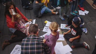A group of children and adults draw on paper on the floor.