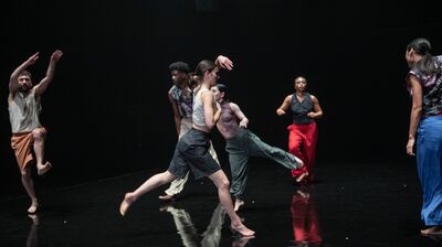 Dancers move across a black floor, one leaping, one with both arms above their head