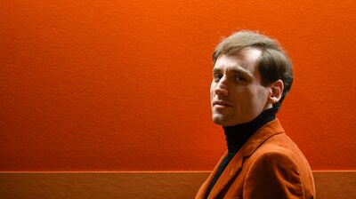 A man wearing an orange cordroy suit jacket, standing in front of an orange wall