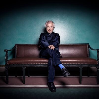 A man wearing a navy suit sat on a brown sofa with his legs crossed and hand resting on his knee