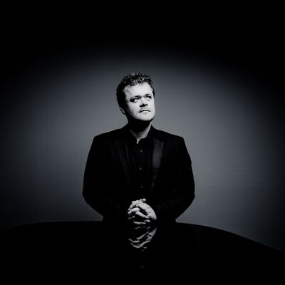 A black and white image of a man standing, with his hands resting on a piano