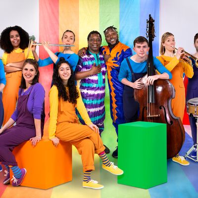 A group of musicians pose together in front of a vibrant rainbow-colored backdrop. They are dressed in bright colours, and are holding various instruments