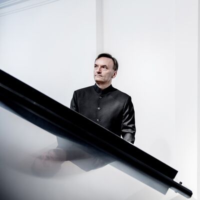 A man wearing a black suit stood behind a grand piano, against a white backdrop