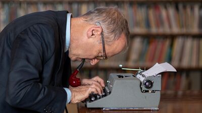 David Sedaris sitting at a typewriter smoking a pipe.