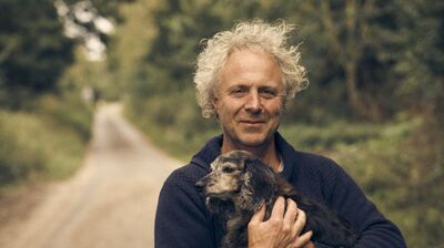 A man with white curly hair standing next to a bike holding a dog on a country lane.