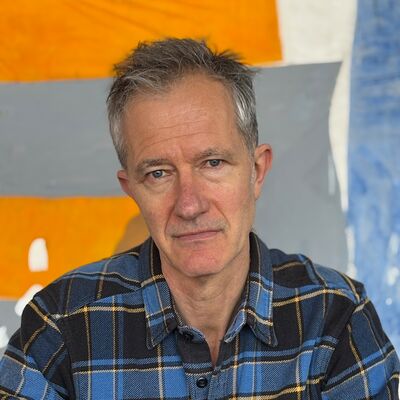 Geoff Dyer in a check shirt against an orange and grey background.