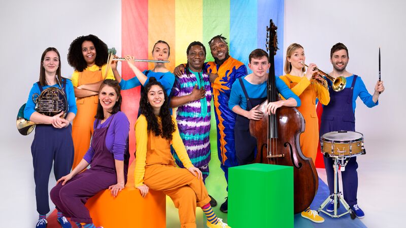 A group of musicians pose together in front of a vibrant rainbow-colored backdrop. They are dressed in bright colours, and are holding various instruments