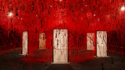 A series of closed doors with keys hanging from the ceiling and red thread attached to the frame connecting it from floor to ceiling creating web like walls