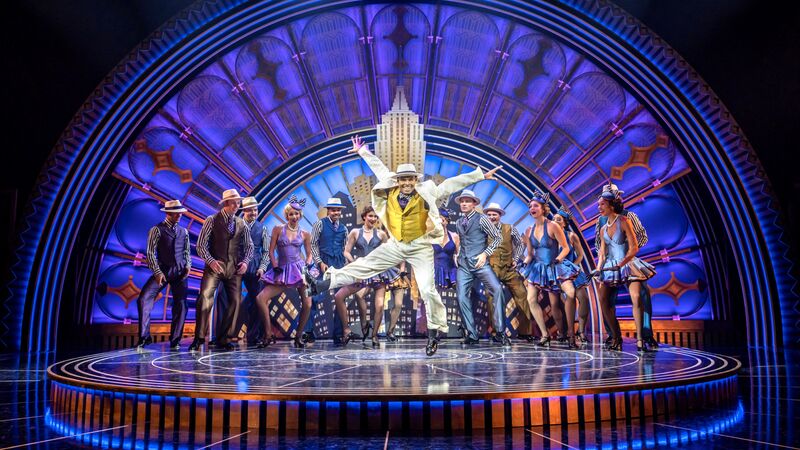 Performers on circular stage wearing short dresses and hats with one in the middle wearing a white suit and leaping in the air