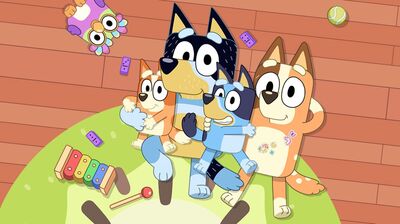 A cartoon dog family, two of which are blue and the other two are brown, lie on a green rug against a wooden floor gazing up at the sky. There is a pink owl toy, a ball and a xylophone on the rug with them.