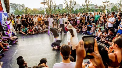 A person doing a head spin on stage with a large crowd seated and stood around the dance battle space