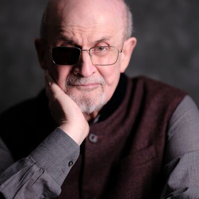 Salman Rushdie wearing a maroon waistcoat and grey shirt with his head in his hand.