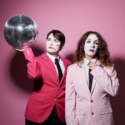 Two people dressed in pink suits with white mime makeup on their faces, one holding a disco ball
