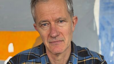 Geoff Dyer in a check shirt against an orange and grey background.