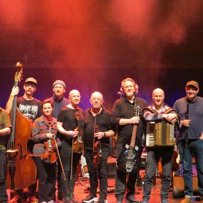 Group of 10 musicians on stage holding a variety of instruments including a double bass, violins, accordion and guitar