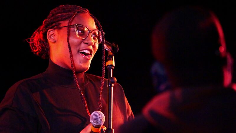 Artist speaking at the microphone at an event wearing glasses and a black turtleneck top
