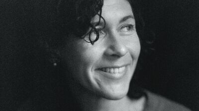 A person with curly hair wearing a button up jumper looking to the right and smiling
