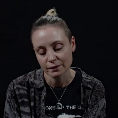 Fran Lock, a woman with her blond hair pulled back and shaved at the sides, looks down whilst reading from their TS Eliot Prize shortlisted collection against a black background