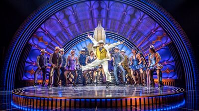 Performers on circular stage wearing short dresses and hats with one in the middle wearing a white suit and leaping in the air