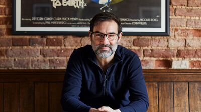 A man with glasses and a beard sits on a wooden bench, smiling in front of a brick wall and vintage poster
