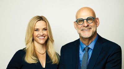 Two people posing for a photo, one with blonde hair and wearing a pantsuit, the other is bald wearing a suit and leaning against a table